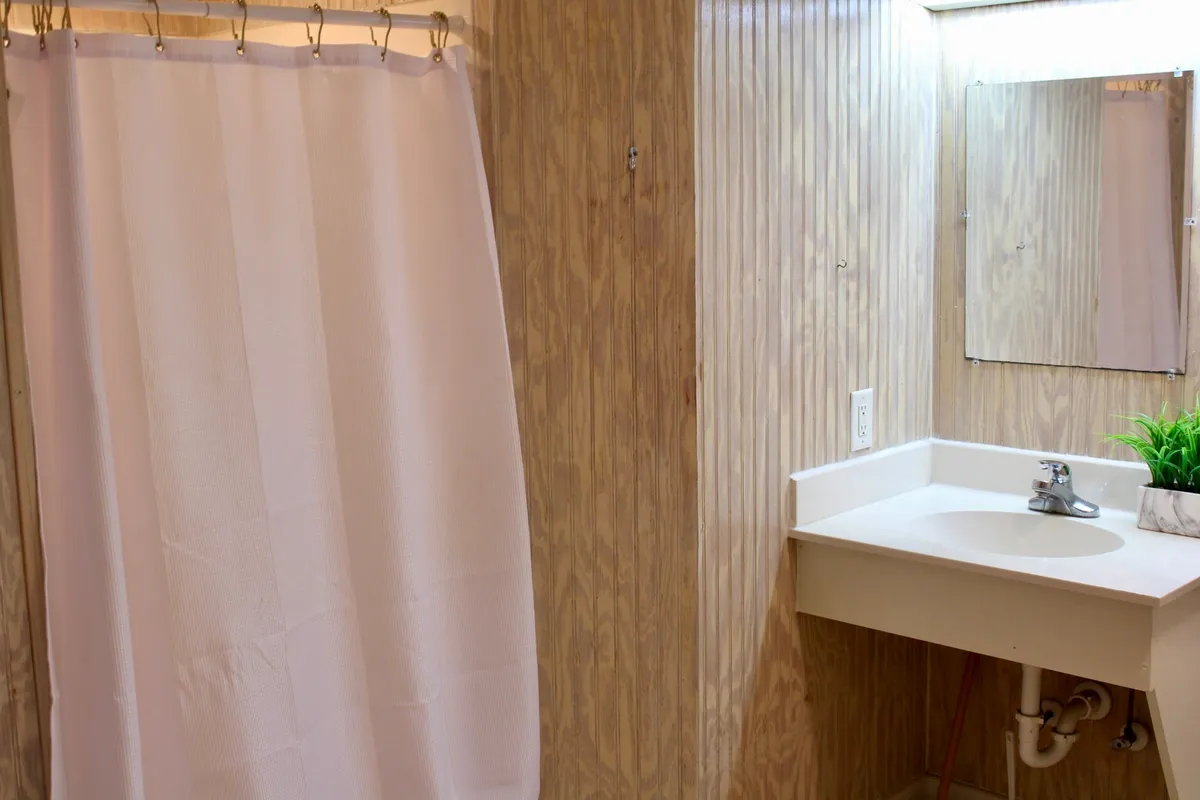 This image showcases a portion of the bathroom featuring a clean shower curtain, a functional sink, and a mirror. The layout is minimalist, with uncluttered surfaces and bright lighting, emphasizing the space's practical and refreshing atmosphere.
