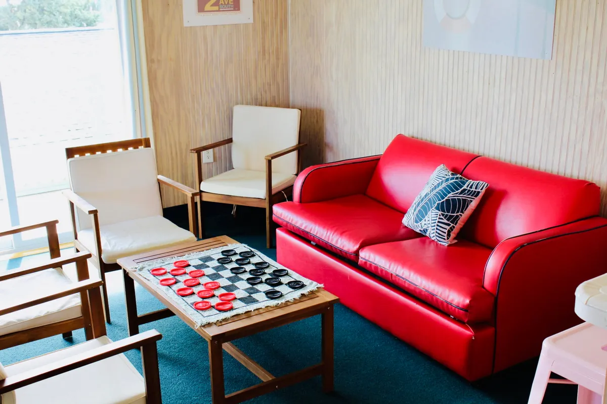 The image showcases a playful and inviting interior space featuring a bold red sofa next to a checkerboard table, surrounded by wooden chairs. The setup suggests a cozy spot for friendly game nights, casual conversations, and relaxation, all bathed in a warm, welcoming atmosphere.