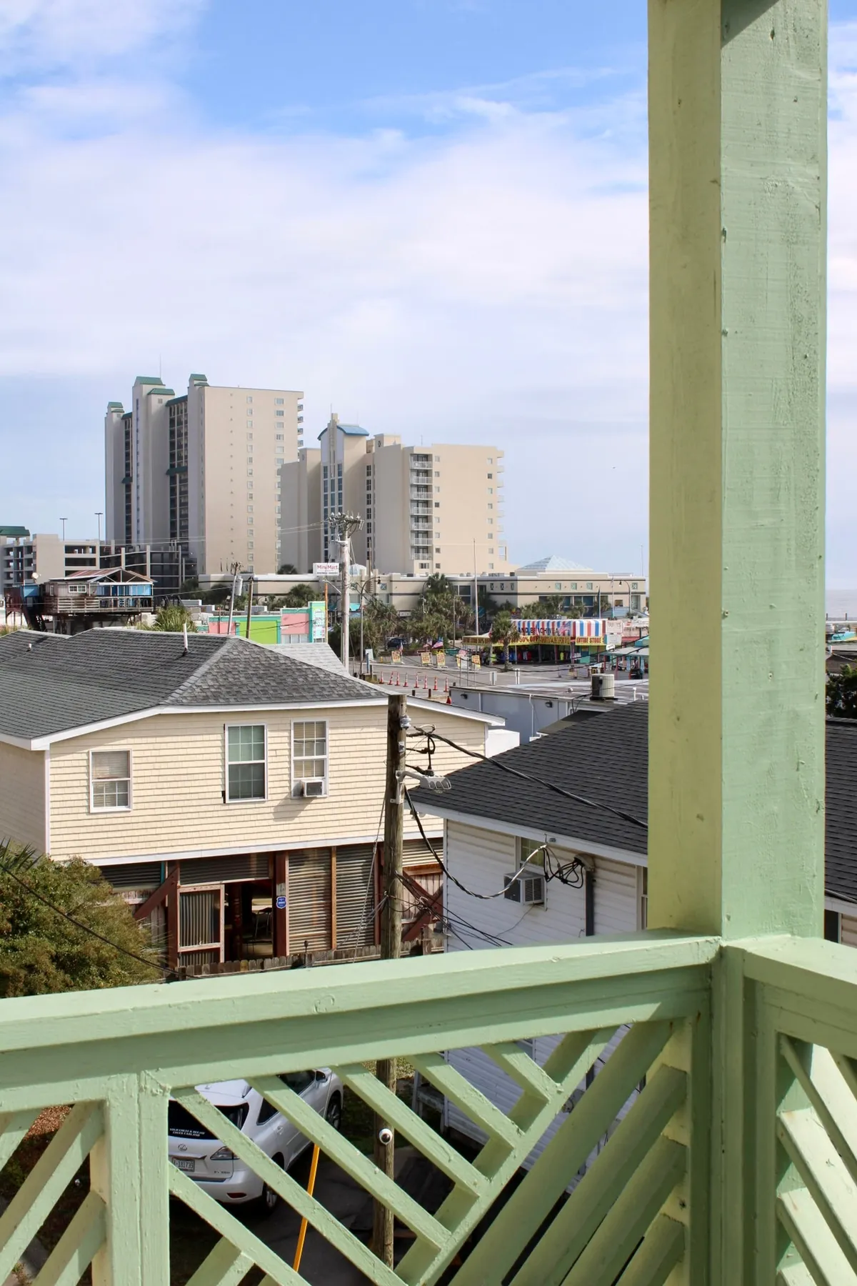 A vibrant balcony scene looking out over beachfront buildings, a lively city skyline, and colorful amusement areas. The green railing frames the view, offering a glimpse of the beach and easy access to outdoor adventures. This setting illustrates the property's close proximity to pet-friendly outdoor spaces for walks and exploration.